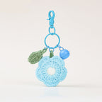 A light blue crocheted flower keychain charm with a white center, attached to a blue plastic clasp and ring. It includes a small green crochet leaf and a light blue bell