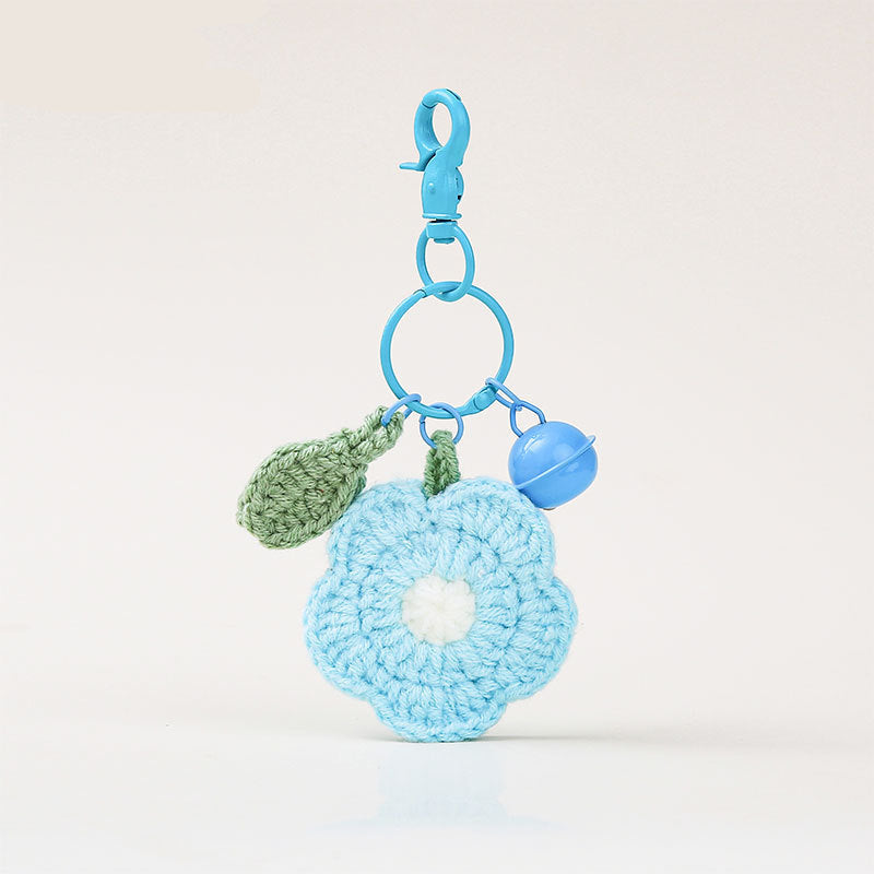 A light blue crocheted flower keychain charm with a white center, attached to a blue plastic clasp and ring. It includes a small green crochet leaf and a light blue bell