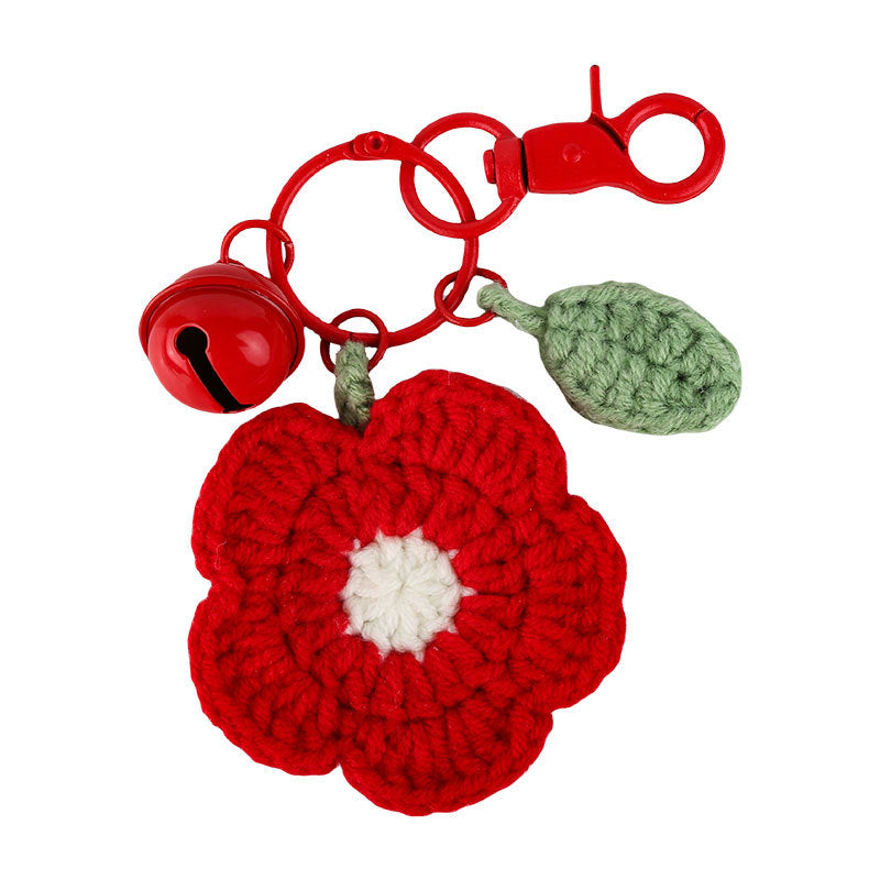 A red crocheted flower-shaped keychain or bag charm with a white center, attached to a red split ring and lobster clasp. A small red bell hangs on one side, and a light green crocheted leaf is attached on the other