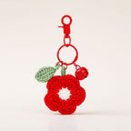 A red crochet flower keychain charm with a white center, green leaf, and small red bell, attached to a red clasp on a light beige background. 