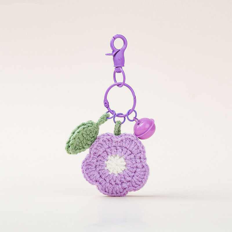 A purple crochet flower-shaped keychain charm with a white center, green leaf, and purple bell, attached to a purple clasp 